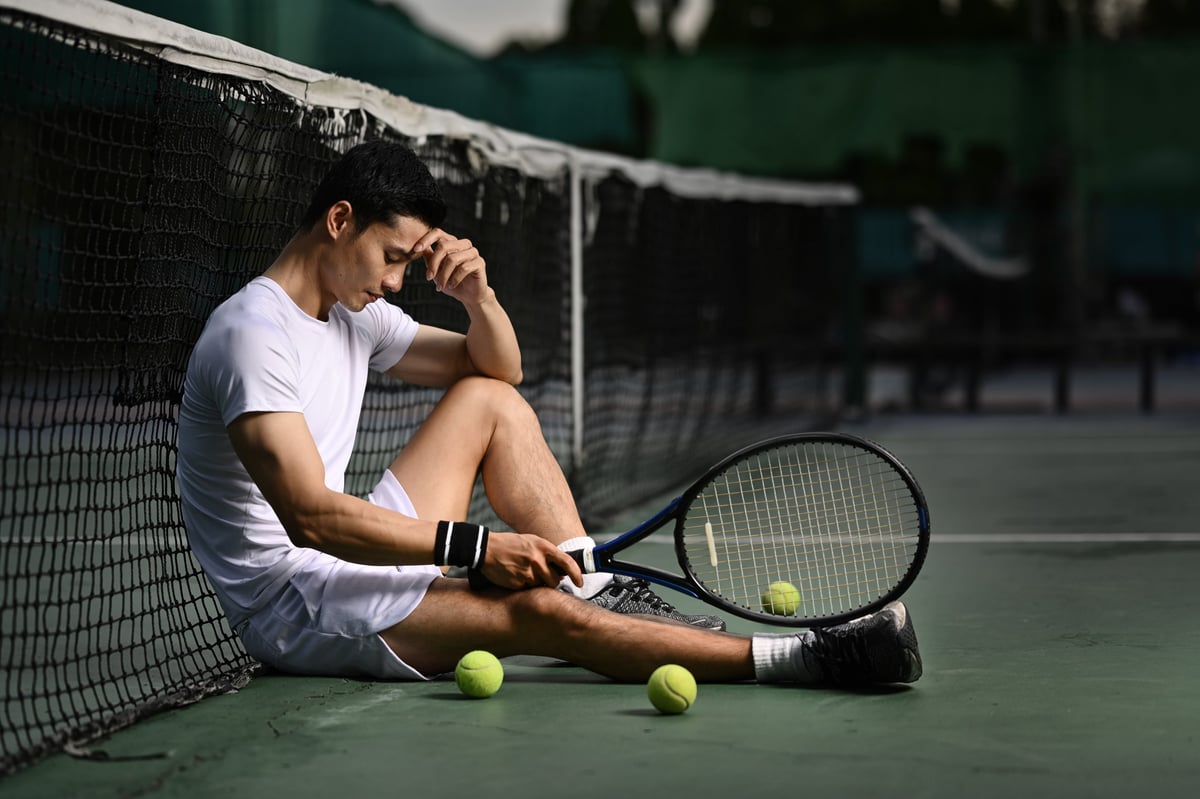 Side view of disappointed asian male tennis player sitting on the court, upset about competition loss, failure emotions.